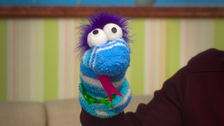 The sockpuppet Chuy. He’s a predominantly blue sockpuppet with white stripes, a green bowtie, a red forked tongue, googly eyes, and purple fuzz for hair.