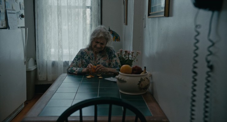Olga, an old woman, sitting at her kitchen table struggling to peel an orange.