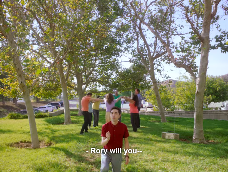 Todd kneeling on a grassy lawn, with a group of dancers behind him in the background, holding a ring, and saying, "Rory, will you," before being cut off.